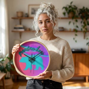 May include: Round wall clock with a wooden frame. The clock face features a teal bear silhouette and the words "grandma bear" in cursive against a pink, purple, and blue background. Black clock hands indicate the time.