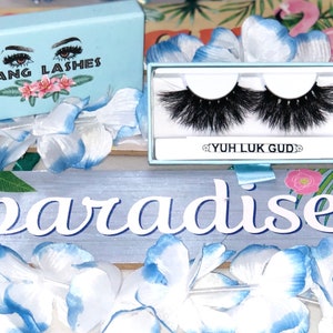 May include: A pair of false eyelashes in a blue box with the text "YUH LUK GUD" on it. The box is surrounded by blue and white flower petals. The box is sitting on a wooden sign that says "paradise".