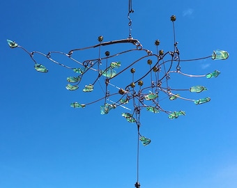 May include: A mobile sculpture with a delicate, intricate design. It features a network of thin wires and numerous small, teardrop-shaped glass pieces in shades of green. The sculpture is suspended from the ceiling and hangs against a clear blue sky.