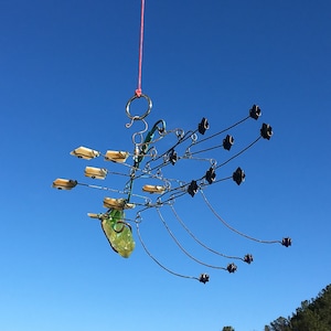 May include: A mobile made of wire and glass hangs from a red string against a blue sky. The mobile features a green glass piece, multiple small white glass pieces, and several black metal pieces.