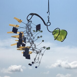 Può includere: Una carillon eolico artigianale con un design complesso. Presenta una varietà di forme e colori, tra cui pezzi di vetro giallo, blu, verde e trasparente, e una grande pietra di vetro verde irregolare. La lavorazione del metallo è intricata e scura.