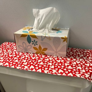 May include: A tissue box with floral designs in shades of blue, yellow, and white sits atop a red and white heart-patterned cover. The tissue box dispenses white tissues. The cover is placed on a white toilet.