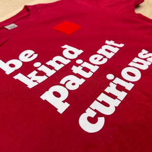 Może przedstawiać: Czerwona koszulka z białym napisem "be kind patient curious". Mały czerwony kwadrat znajduje się nad słowem "kind". Koszulka jest wykonana z miękkiego materiału.