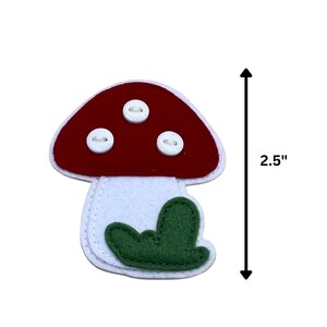 Może przedstawiać: Filcowa aplikacja w kształcie grzyba, o wysokości około 6,35 cm. Grzyb ma czerwony kapelusz z trzema białymi guzikami, biały trzon i zielony detal liścia. Idealny do rękodzieła i szycia.