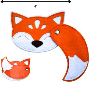 Könnte beinhalten: Ein Fuchspielzeug aus Filz mit orangefarbenem Körper und weißen Akzenten. Der Fuchskopf ist 10 cm breit. Der Fuchs hat eine schwarze Nase und ein lächelndes Gesicht. Der Schwanz ist mit einer Metallniete befestigt. Ein kleinerer Fuchskopf ist ebenfalls enthalten.