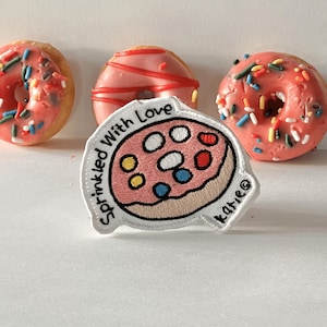 Può includere: Toppa ricamata bianca con un donut rosa con codette colorate e il testo "Sprinkled With Love" e "katie" in un cerchio bianco.