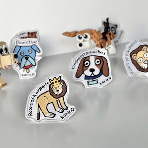 May include: A set of embroidered iron-on patches featuring various animals with positive messages. The patches are white with colorful embroidery. The patches include a dog with the words "Pawsitive", a lion with the words "Roars of Kindness", a dog with the words "Kindness Unleashed", and a lion with a crown and the words "Roars of Kindness".