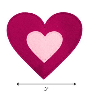 Puede incluir: Un corazón de fieltro magenta con un corazón rosa claro en el centro. El corazón de fieltro mide 7,6 cm de ancho. La imagen es una foto de cerca del corazón, probablemente un artículo de artesanía o decoración.