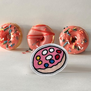 Può includere: Una toppa ricamata a forma di ciambella rosa con codette colorate. La toppa è circondata da tre ciambelle rosa con diverse guarnizioni.