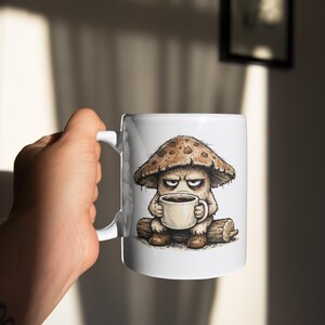 May include: White ceramic coffee mug featuring a grumpy mushroom character with a coffee cup. The mushroom is brown and tan, with an angry expression. The mug has a white handle and is held by a hand.