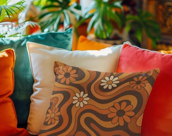 May include: Decorative throw pillows in shades of orange, teal, and cream. The focal point is a pillow with a brown, orange, and cream daisy pattern. The pillows are arranged on a burnt orange sofa, with lush green plants in the background.