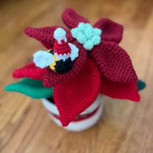 May include: A handmade crochet poinsettia flower in a white pot with a red stripe. The flower is a deep red color with green leaves and a light green center. A crochet bee wearing a Santa hat is attached to the flower.