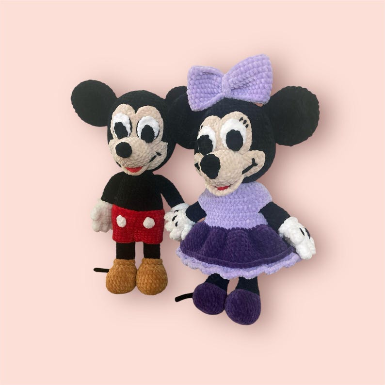 May include: A crocheted plush toy set featuring Mickey Mouse and Minnie Mouse. Mickey is wearing red shorts and brown shoes. Minnie is wearing a purple dress with a lavender bow and purple shoes.