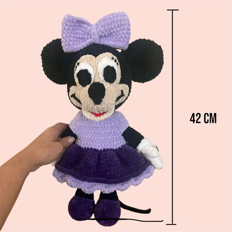 May include: A crocheted plush toy of a purple-dressed Mickey Mouse character, standing 42 cm tall. The toy has a purple dress with a white bow, black ears, and a black tail.