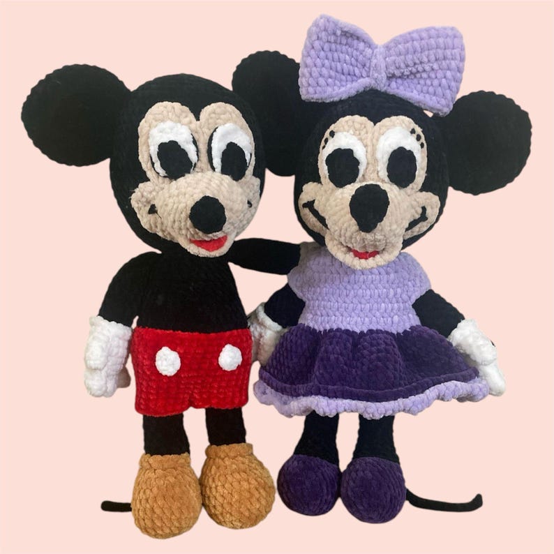 May include: A crocheted Mickey Mouse and Minnie Mouse plush toy set. Mickey is wearing red shorts with white polka dots and Minnie is wearing a purple dress with a lavender bow.