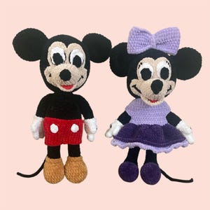 May include: A pair of crocheted Mickey Mouse and Minnie Mouse plush toys. Mickey is wearing red shorts and brown shoes. Minnie is wearing a purple dress and purple shoes. Both have black eyes and white accents.