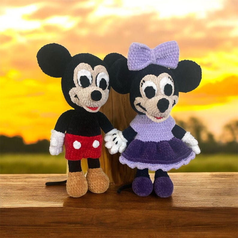 May include: A crocheted Mickey Mouse and Minnie Mouse plush toy set. Mickey is wearing red shorts with white polka dots and Minnie is wearing a purple dress with a white bow.