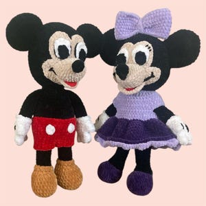 May include: A crocheted Mickey Mouse and Minnie Mouse plush toy set. Mickey is wearing red shorts with white polka dots and brown shoes. Minnie is wearing a purple dress with a purple bow and purple shoes.