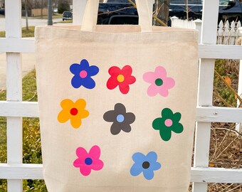 Peut inclure: Un sac fourre-tout en toile blanche avec des motifs de fleurs colorées. Les fleurs sont dans des tons de bleu, rose, rouge, jaune et vert.