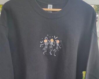 May include: Black sweatshirt with an embroidered design of three skeletons dancing with pumpkin heads. The skeletons are white and the pumpkins are orange.