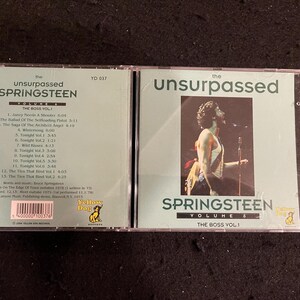 Puede incluir: Dos estuches de CD con los álbumes "The Unsurpassed Springsteen" de Bruce Springsteen, Volumen 1 y Volumen 6. Las portadas de los álbumes muestran texto y una foto de Springsteen actuando. Los estuches de CD están en una funda de plástico transparente.