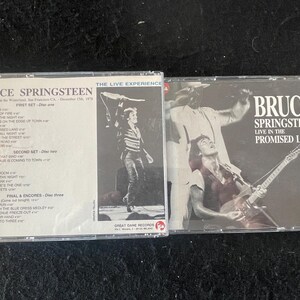 Może przedstawiać: Dwie kasety magnetofonowe z albumami Bruce'a Springsteena. Pierwsza kaseta to "The Live Experience" i zawiera czarno-białe zdjęcie Bruce'a Springsteena na scenie. Druga kaseta to "Live in the Promised Land" i zawiera kolorowe zdjęcie Bruce'a Springsteena grającego na gitarze na scenie.