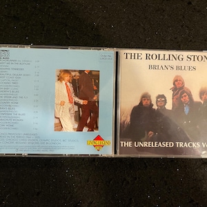 Könnte beinhalten: Zwei CDs mit The Rolling Stones. Auf einem steht "Brian's Blues" und "The Unreleased Tracks Vol. 2". Das andere hat eine Titelliste und ein Foto einer Person. Die CDs befinden sich in transparenten Hüllen.