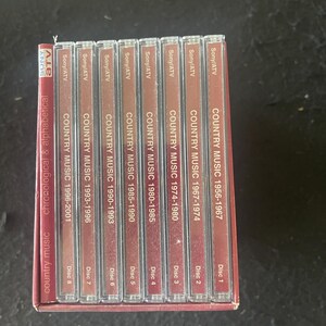 May include: A set of eight CDs containing a compilation of country music from 1956 to 2001. The CDs are labeled with the years and the disc number. The set is in a red and white box with the title "Country Music Encyclopedia" on the front.