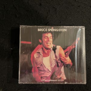 Puede incluir: Una portada de casete con Bruce Springsteen, un músico, tocando la guitarra. La portada es roja y negra con el texto "Bruce Springsteen" y "Live and unreleased 1971/79".