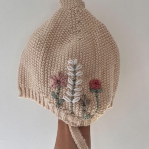 May include: A beige knitted bonnet with embroidered floral details. The bonnet features pink, white, and green flowers. A knitted tie is attached. The bonnet is made of a soft, textured material.