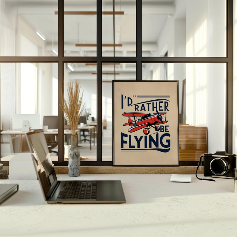 Op de afbeelding: Een ingelijste poster met een illustratie van een rood en wit vliegtuig en de tekst "I'd rather be flying". De poster hangt aan een muur in een modern kantoor.