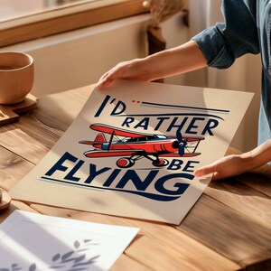 Op de afbeelding: Een beige poster met de tekst "I'd rather be flying" in blauwe en rode letters. Een rood en wit vliegtuig vliegt boven de tekst.