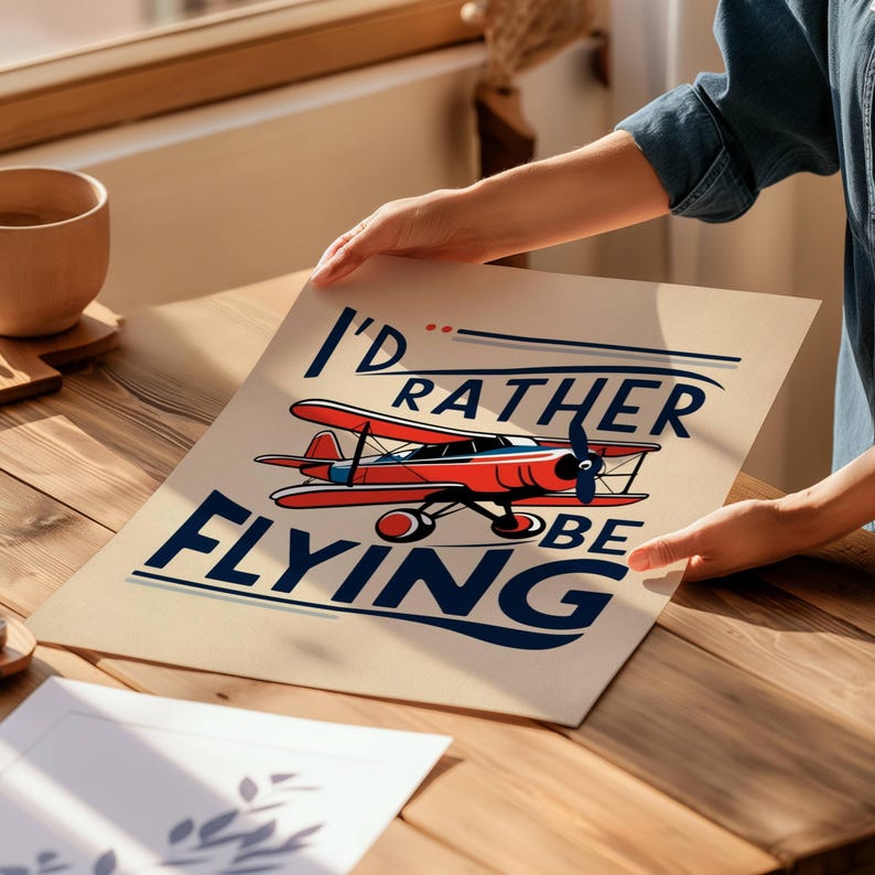 Op de afbeelding: Een beige poster met de tekst "I'd rather be flying" in blauwe en rode letters. Een rood en wit vliegtuig vliegt boven de tekst.