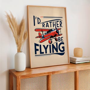 Op de afbeelding: Een vintage poster met een rood vliegtuig en de tekst "I'd rather be flying". De poster heeft een beige achtergrond met blauwe en witte accenten.