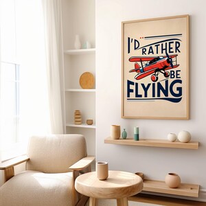 Op de afbeelding: Een vintage poster met een rood vliegtuig en de tekst "I'd rather be flying". De poster is ingelijst met hout en hangt aan een witte muur.