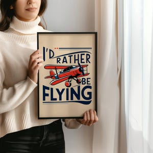 Op de afbeelding: Een ingelijst print met een vintage ontwerp met een rood vliegtuig en de tekst "I'd rather be flying".