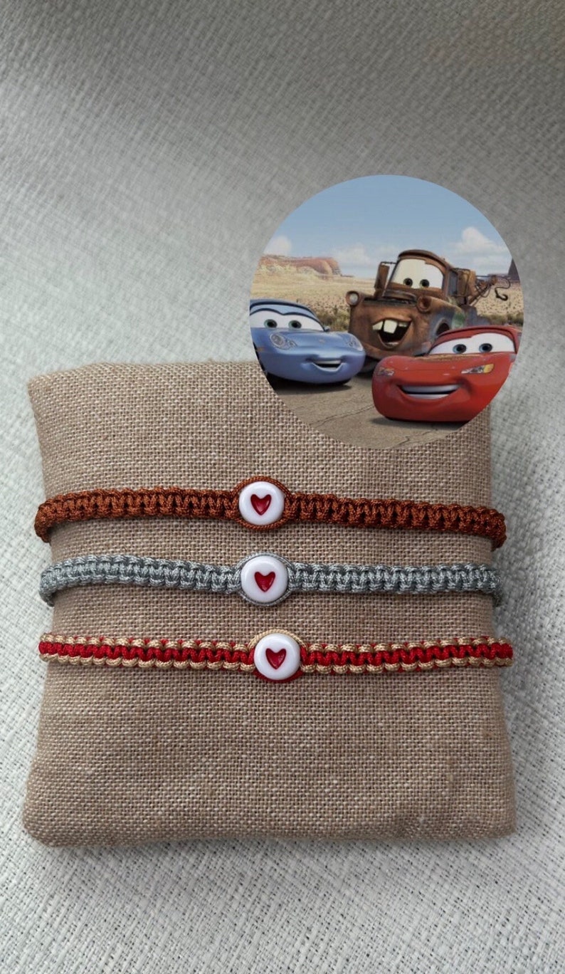 May include: Three friendship bracelets with red, brown and grey cords and white heart-shaped buttons. The bracelets are displayed on a brown fabric surface. The background image shows three cartoon cars from the movie Cars.