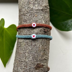 May include: Two friendship bracelets with heart charms. One bracelet is brown and the other is light blue.