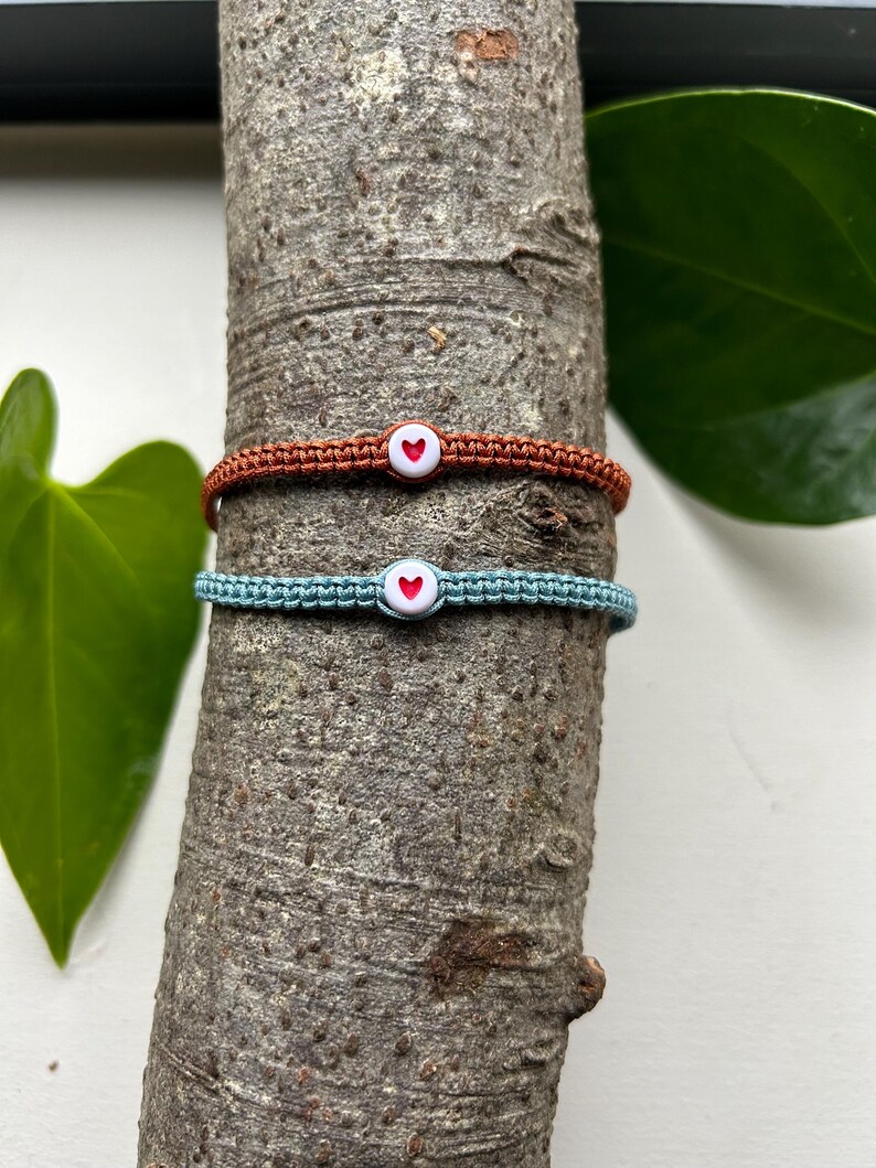 May include: Two friendship bracelets with heart charms. One bracelet is brown and the other is light blue.