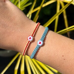 May include: Two friendship bracelets with heart-shaped buttons. One bracelet is red and yellow, the other is blue.