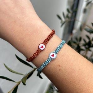 May include: Two friendship bracelets with heart-shaped buttons. One bracelet is light blue and the other is dark orange.