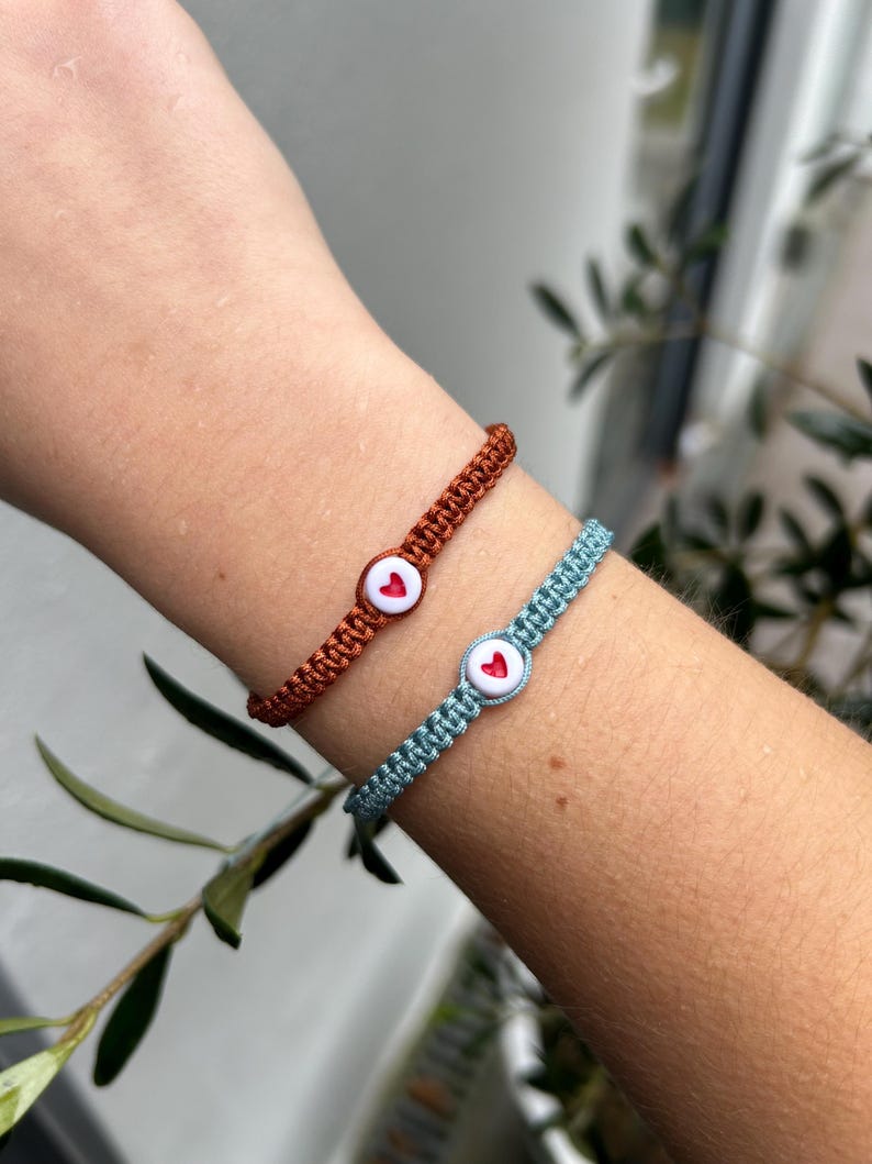 May include: Two friendship bracelets with heart-shaped buttons. One bracelet is light blue and the other is dark orange.