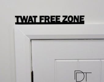 May include: Black door sign with the text "TWAT FREE ZONE" in all caps. The sign is mounted above a white door with a black "DT" logo and the text "DOOR TOPPERS" below.