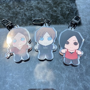 May include: Three acrylic keychains with cartoon character designs. Each keychain has a different character with a black metal clasp. The characters are wearing different outfits and have large eyes. One keychain has the letters "R.P.D." on the character's outfit.