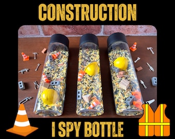 Puede incluir: Tres botellas de vidrio transparente llenas de arroz amarillo y negro y varios juguetes con temática de construcción, incluyendo cascos de obra en miniatura, conos y herramientas. Las botellas están etiquetadas como "I SPY BOTTLE" y son una actividad sensorial divertida para los niños.