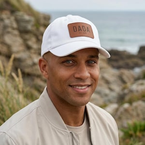 May include: A white baseball cap featuring a rectangular brown leather patch on the front. The patch is embossed with the text DADA and the year EST. 2025 in a dark font. The cap has a structured crown and a curved brim, providing a clean look.