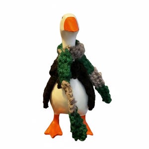 May include: A white goose figurine with an orange beak and orange webbed feet. The goose is wearing a handmade crocheted scarf featuring a color pattern of dark green, black, and light gray yarn, wrapped around its neck and hanging down.