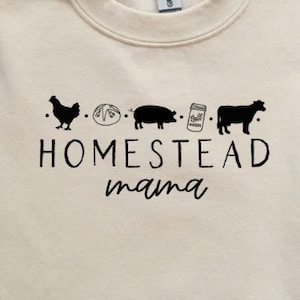 Op de afbeelding: Een cr&egrave;mekleurig sweatshirt met een zwart grafisch ontwerp met de tekst "Homestead mama" met een rij pictogrammen boven de tekst: een kip, een brood, een varken, een pot met een deksel en een koe.