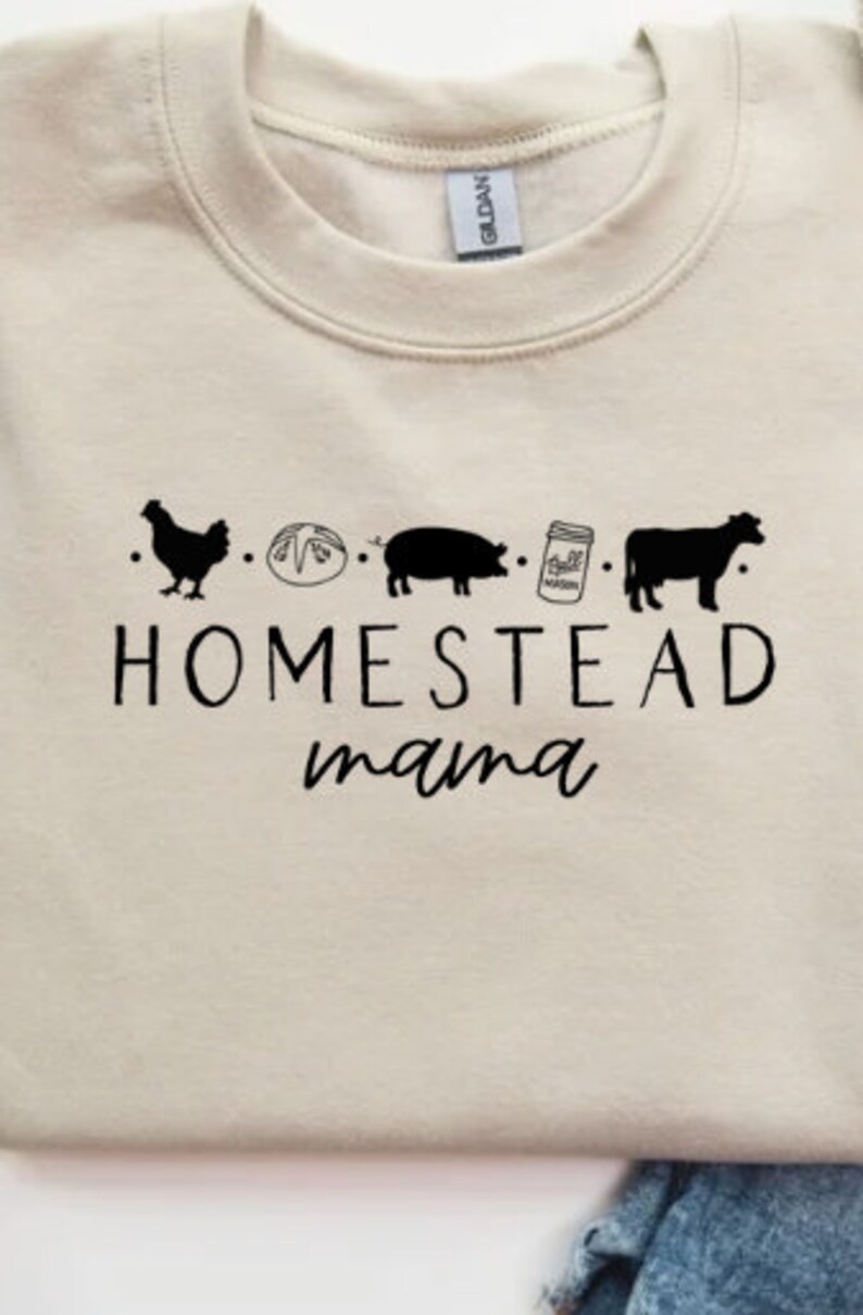 Op de afbeelding: Een cr&egrave;mekleurig sweatshirt met een zwart grafisch ontwerp met de tekst "Homestead mama" met een rij pictogrammen boven de tekst: een kip, een brood, een varken, een pot met een deksel en een koe.