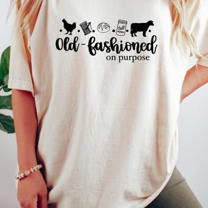 May include: A white t-shirt with a black graphic design that says "Old-fashioned on purpose". The graphic includes a chicken, a spool of thread, a loaf of bread, a mason jar, and a cow.